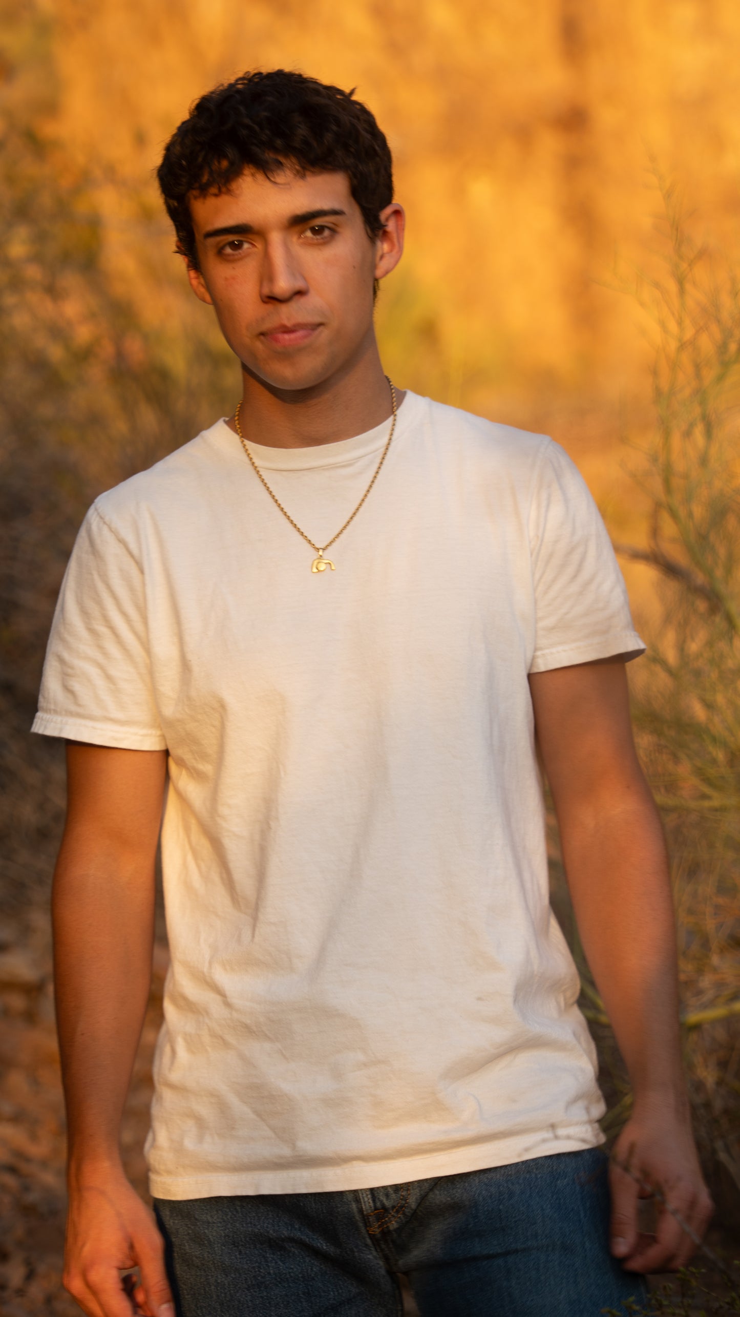 Man wearing a white t-shirt, blue jeans, and empty tomb necklace against a warm-toned natural background